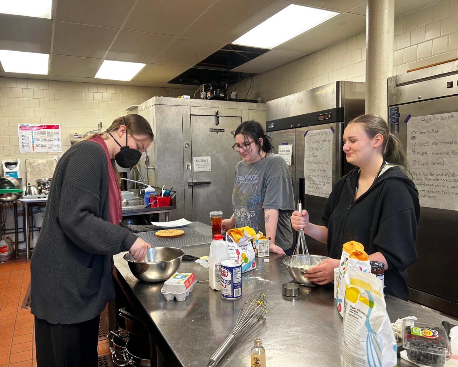Three students measuring ingredients and whisking batter in a kitchen during a culinary class