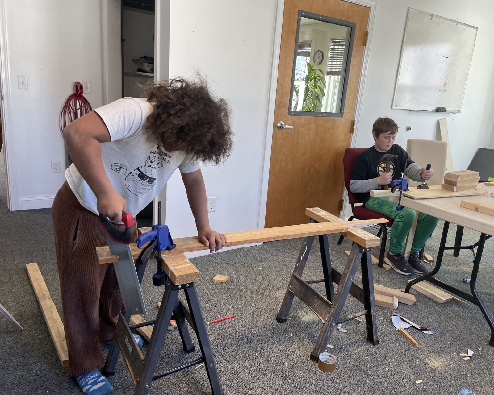 A student sawing a board while another hammers a project together in the woodshop